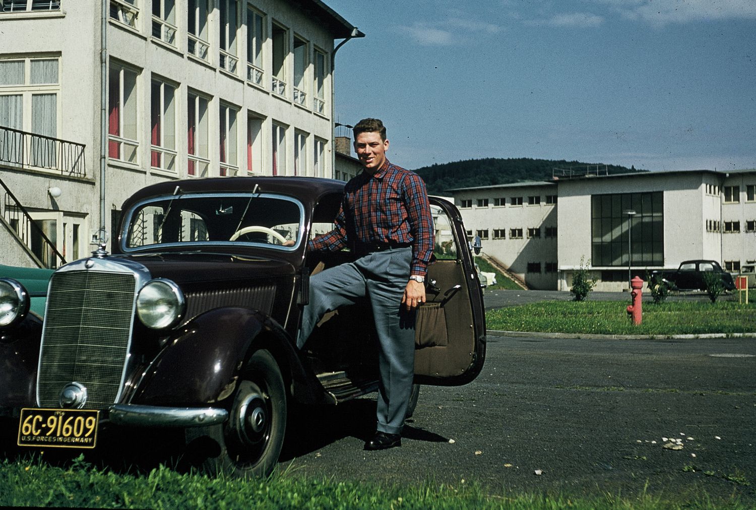 Man leaning against a vintage 1950s Mercedes-Benz car parked outside a European building.