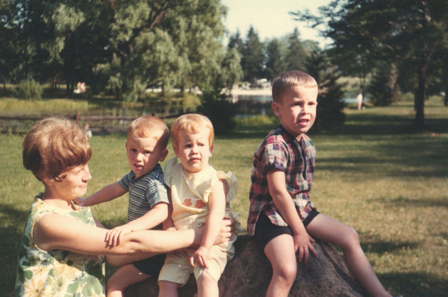 A woman sits outdoors with three young children on a rock in a sunny green park.