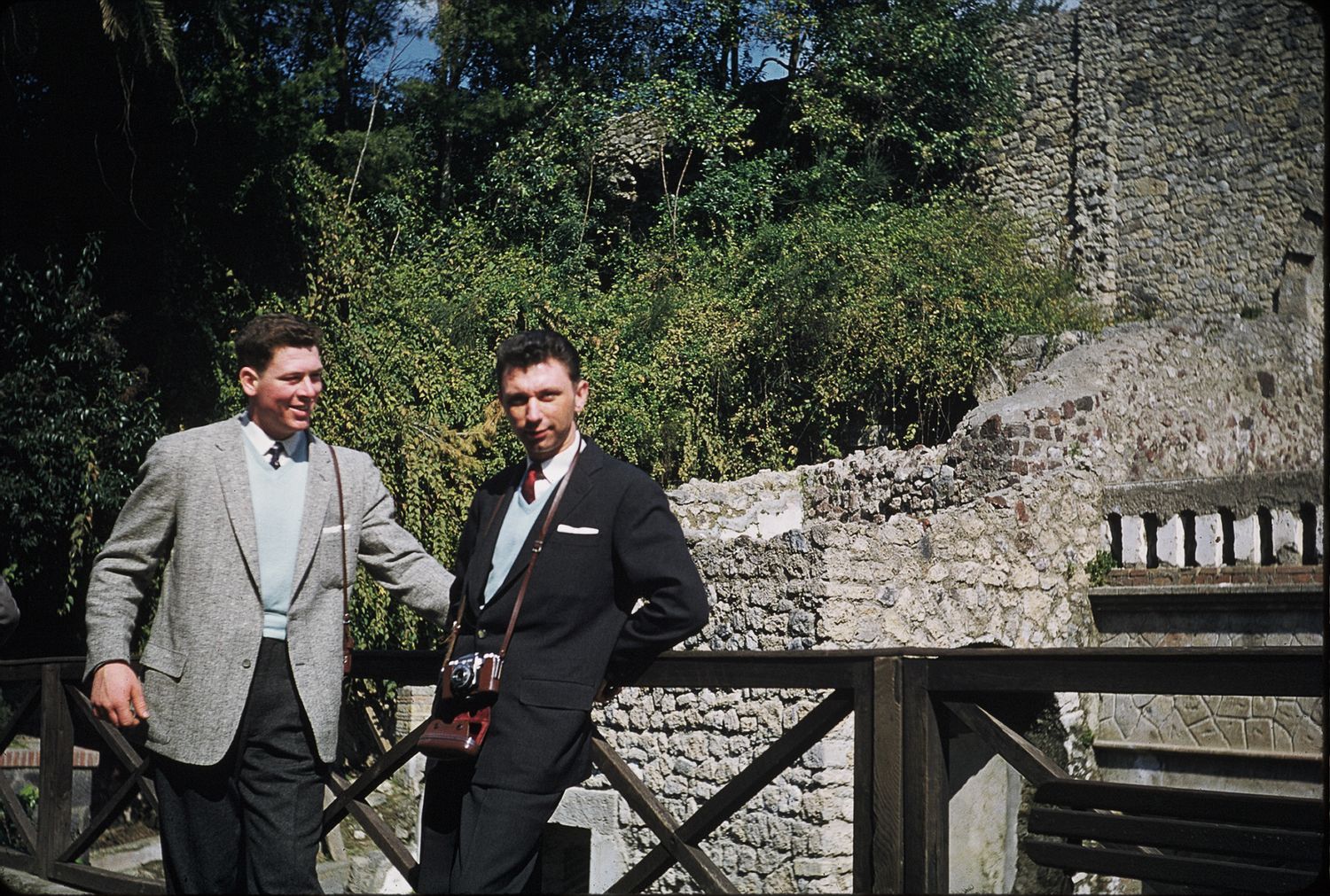 Two men in suits smiling on a wooden bridge near ancient stone ruins outdoors.