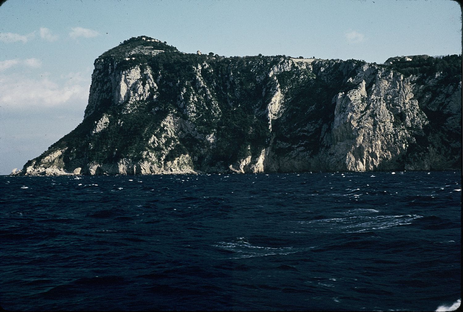 Dramatic rocky cliff face rising from deep blue ocean waters viewed from a boat on a clear day.