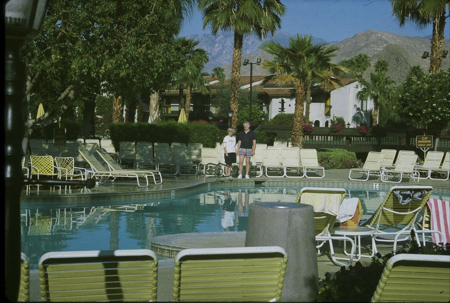 Two people stand beside a sparkling resort swimming pool surrounded by lounge chairs and palm trees.
