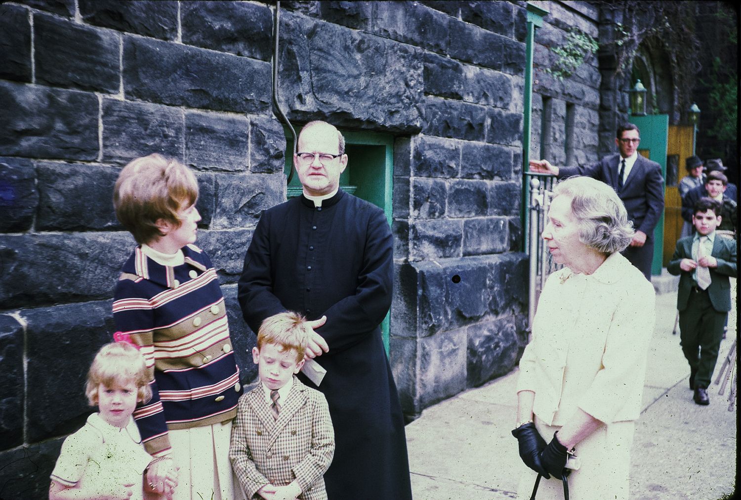 Priest in black cassock greeting elderly woman and family with children outside stone church building.