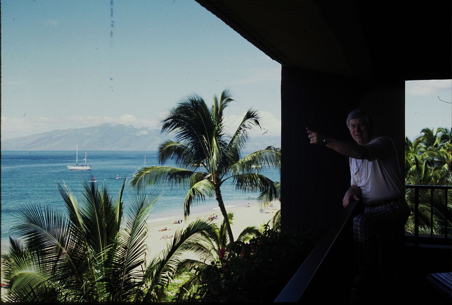A man stands on a tropical balcony overlooking a palm-lined beach with a sailboat on turquoise water.