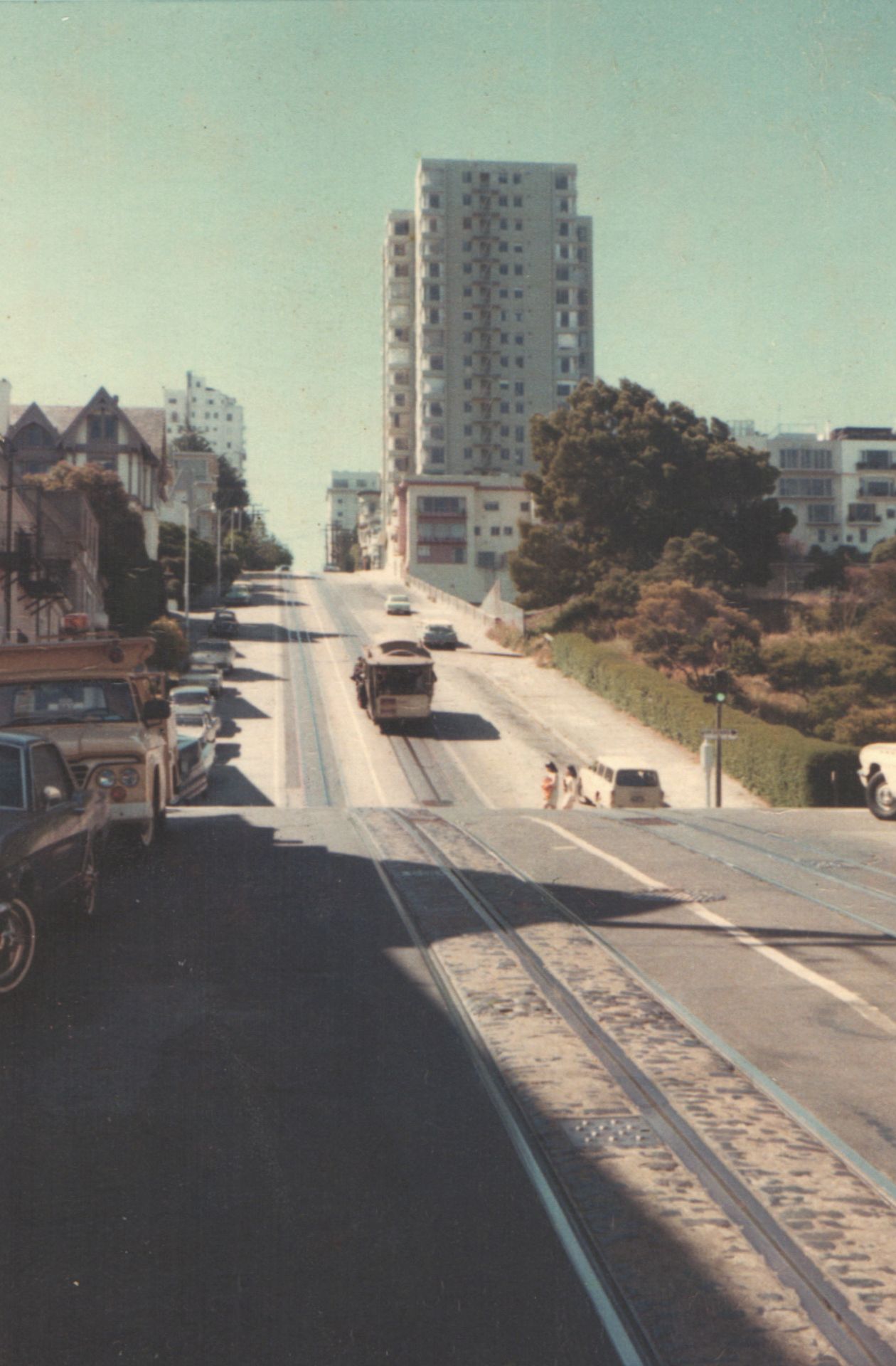 A steep San Francisco street with cable car tracks, parked cars, and a tall white building in the background on a sunny day.