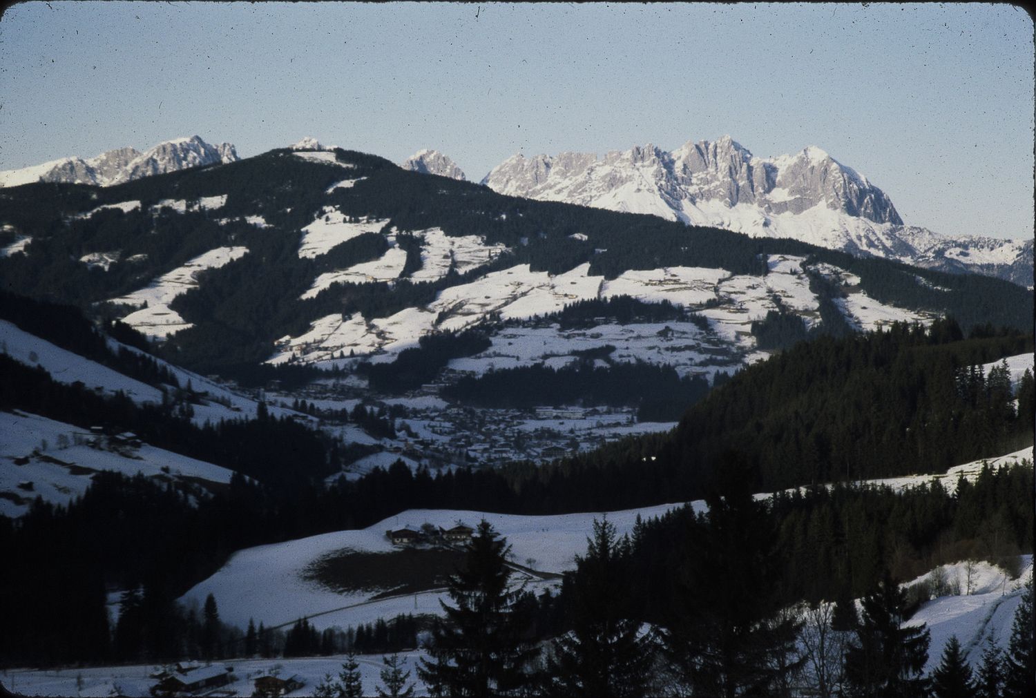 Snow-covered alpine valley surrounded by evergreen forests and dramatic rocky mountain peaks under a clear winter sky.