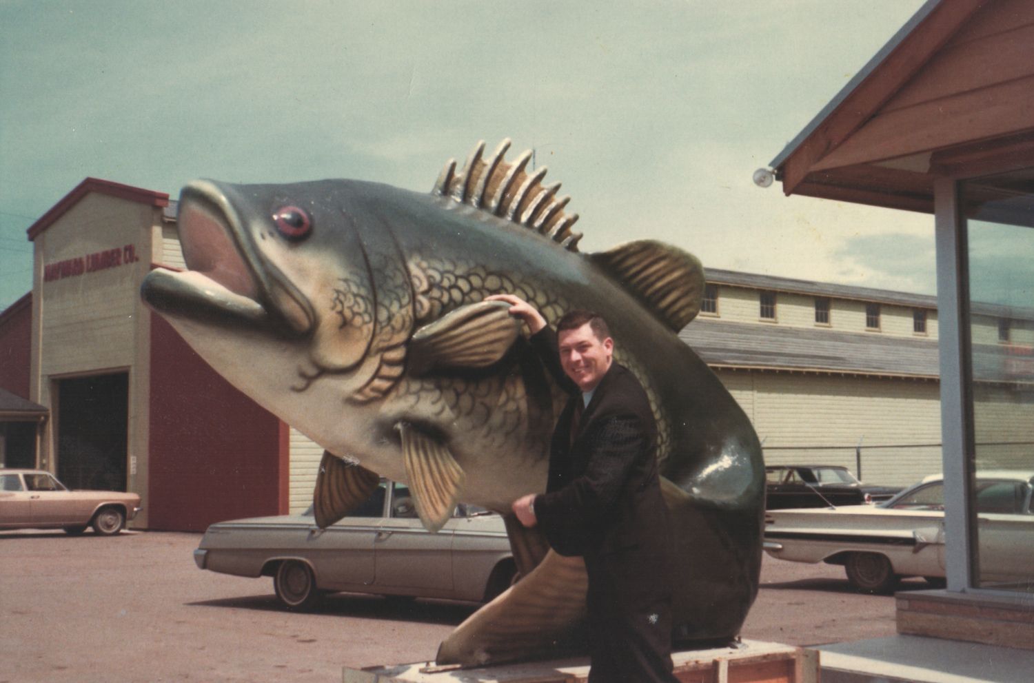 Man posing with a giant largemouth bass sculpture outside a building with vintage cars in the background.