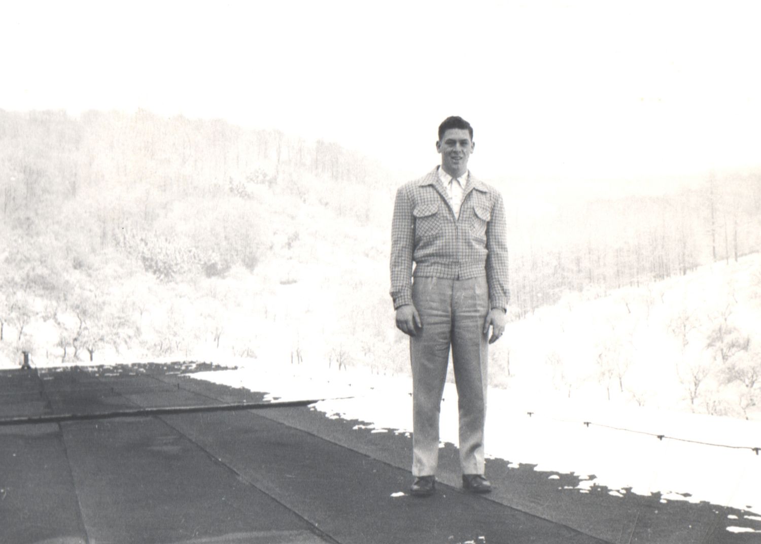 Vintage black and white photo of a man in military uniform standing on a road with snow-covered hills behind him.