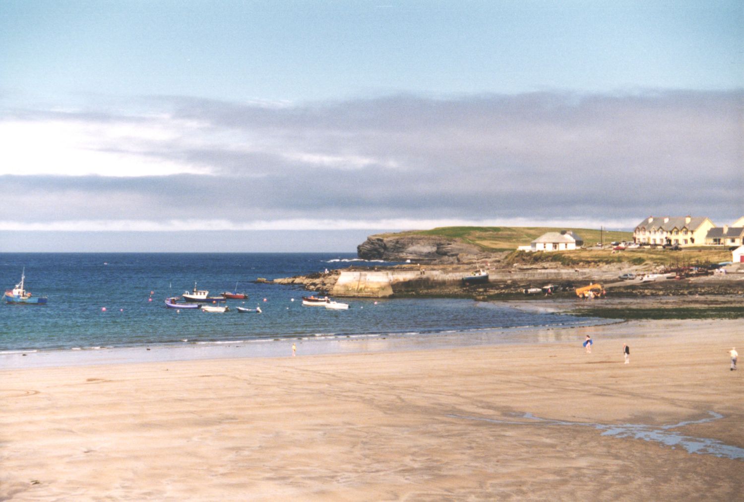 Sandy beach with calm blue waters, moored sailboats, and a small coastal village with white houses under a cloudy sky.