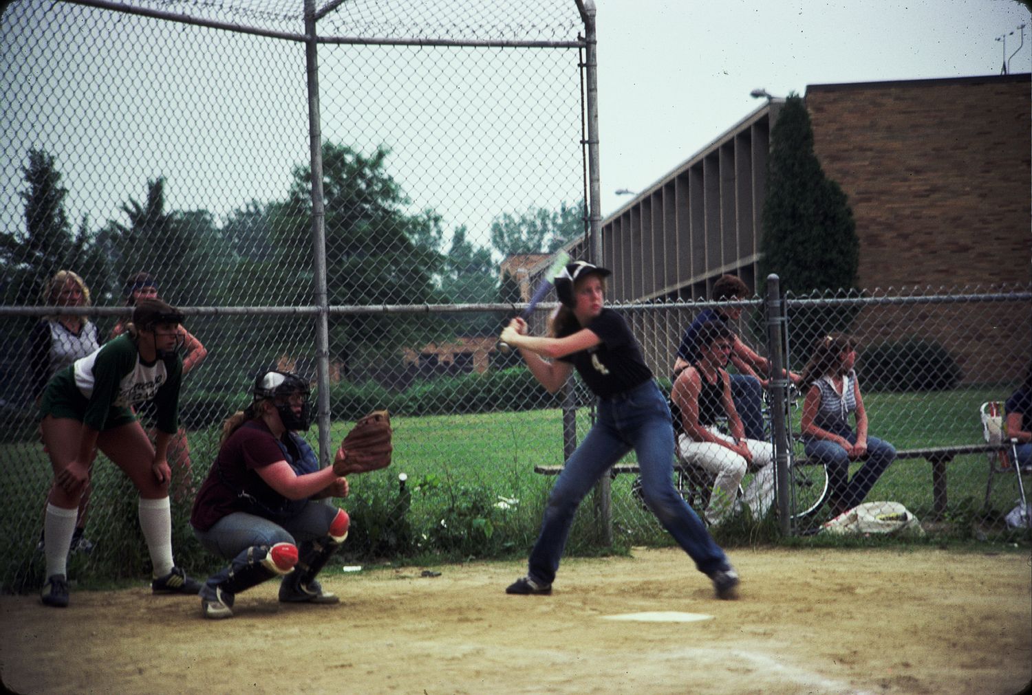 Players compete in a softball game on a sunny day at an outdoor field with a chain-link fence.