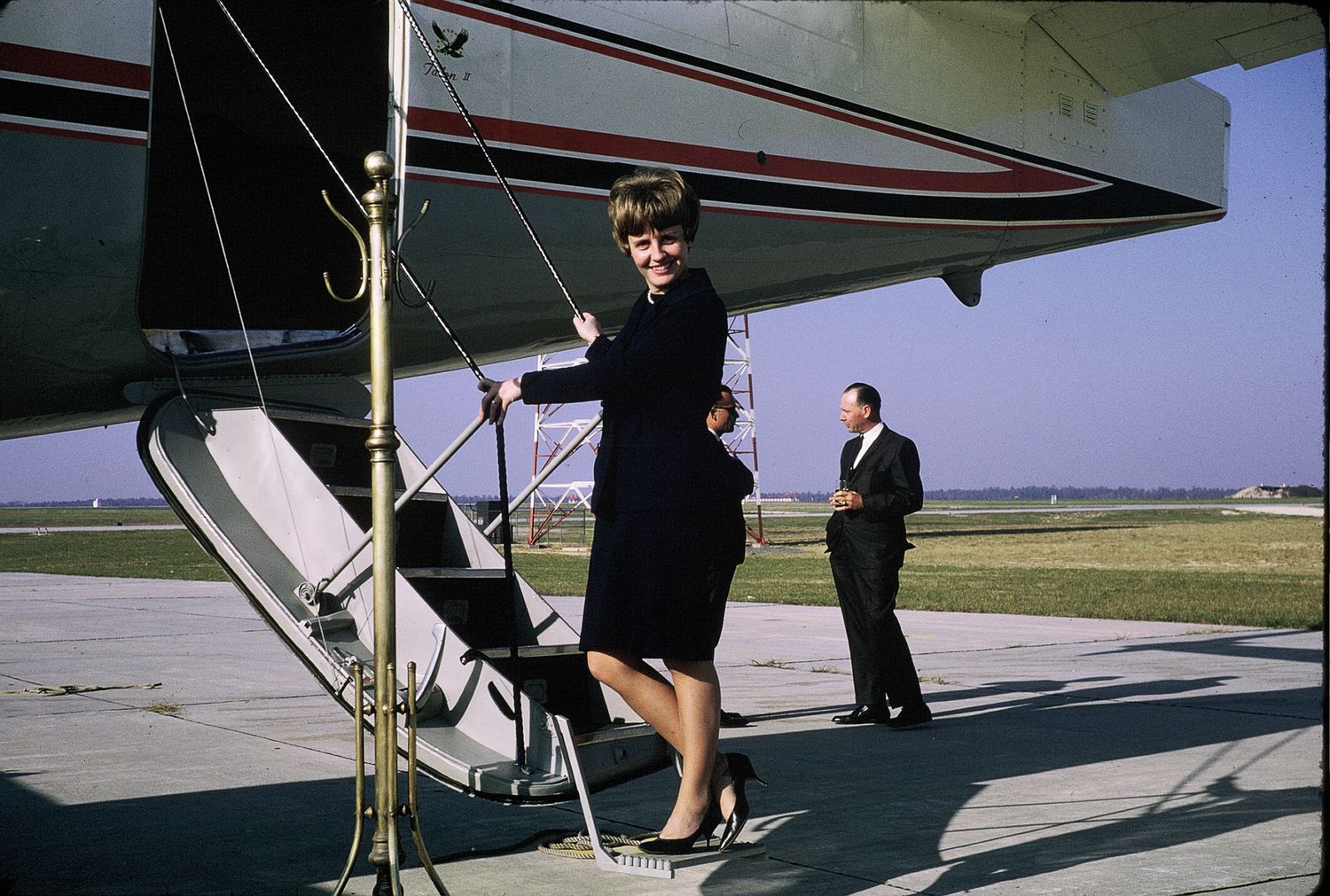Elegant woman in navy suit boarding a vintage propeller aircraft on a sunny airport tarmac.