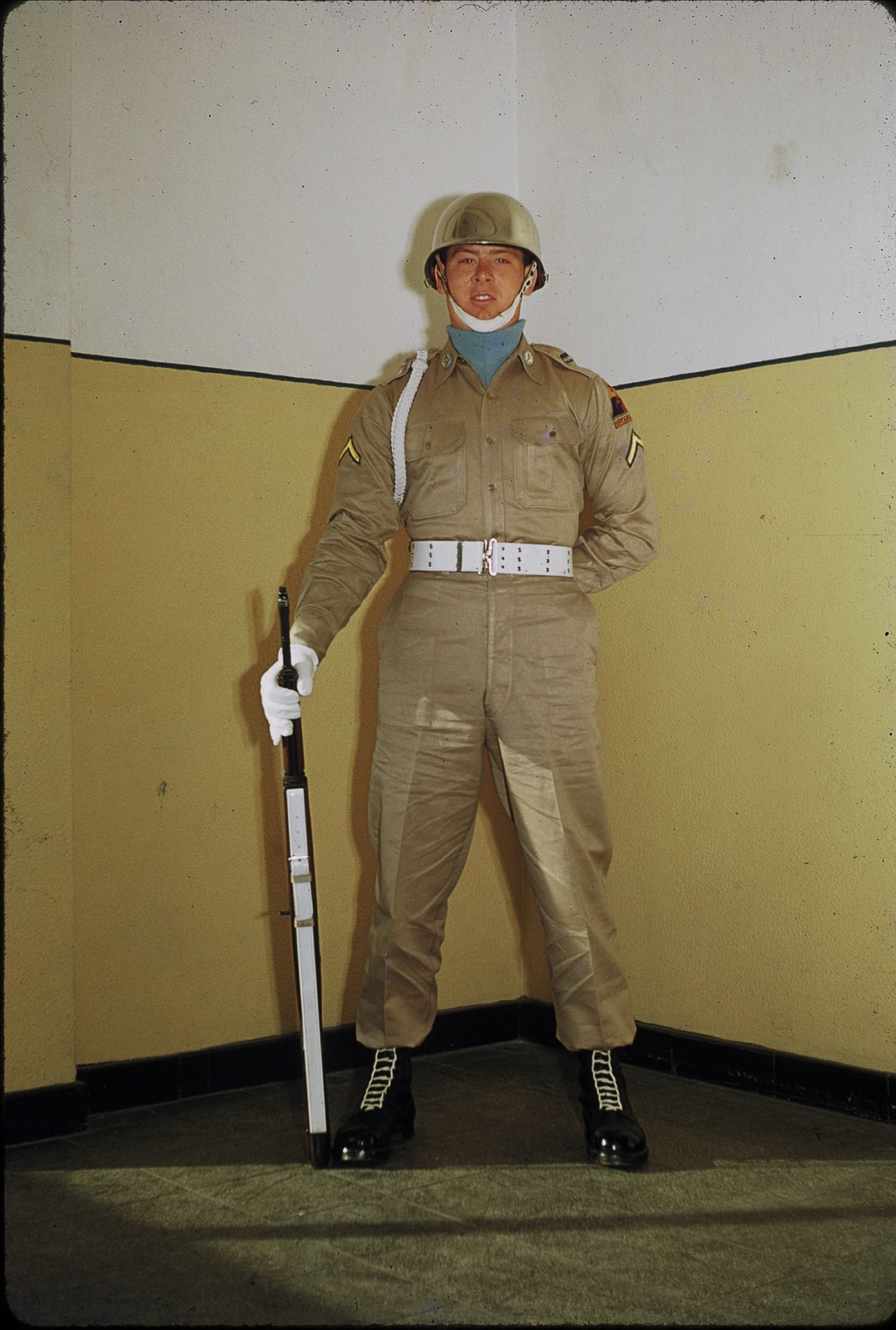 US Army soldier in tan duty uniform standing at attention holding a baton indoors.