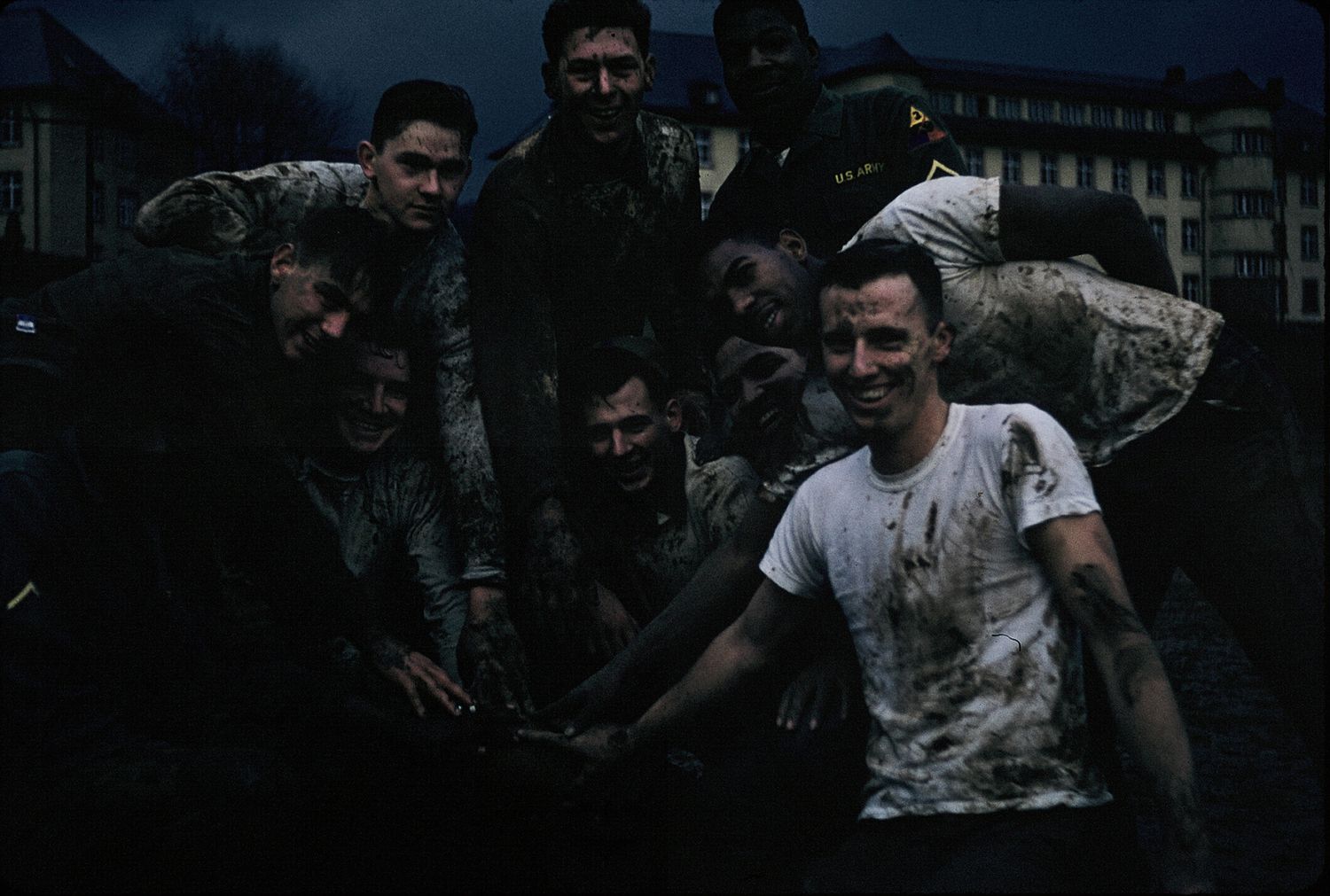 Group of muddy soldiers smiling together outdoors near buildings at dusk.