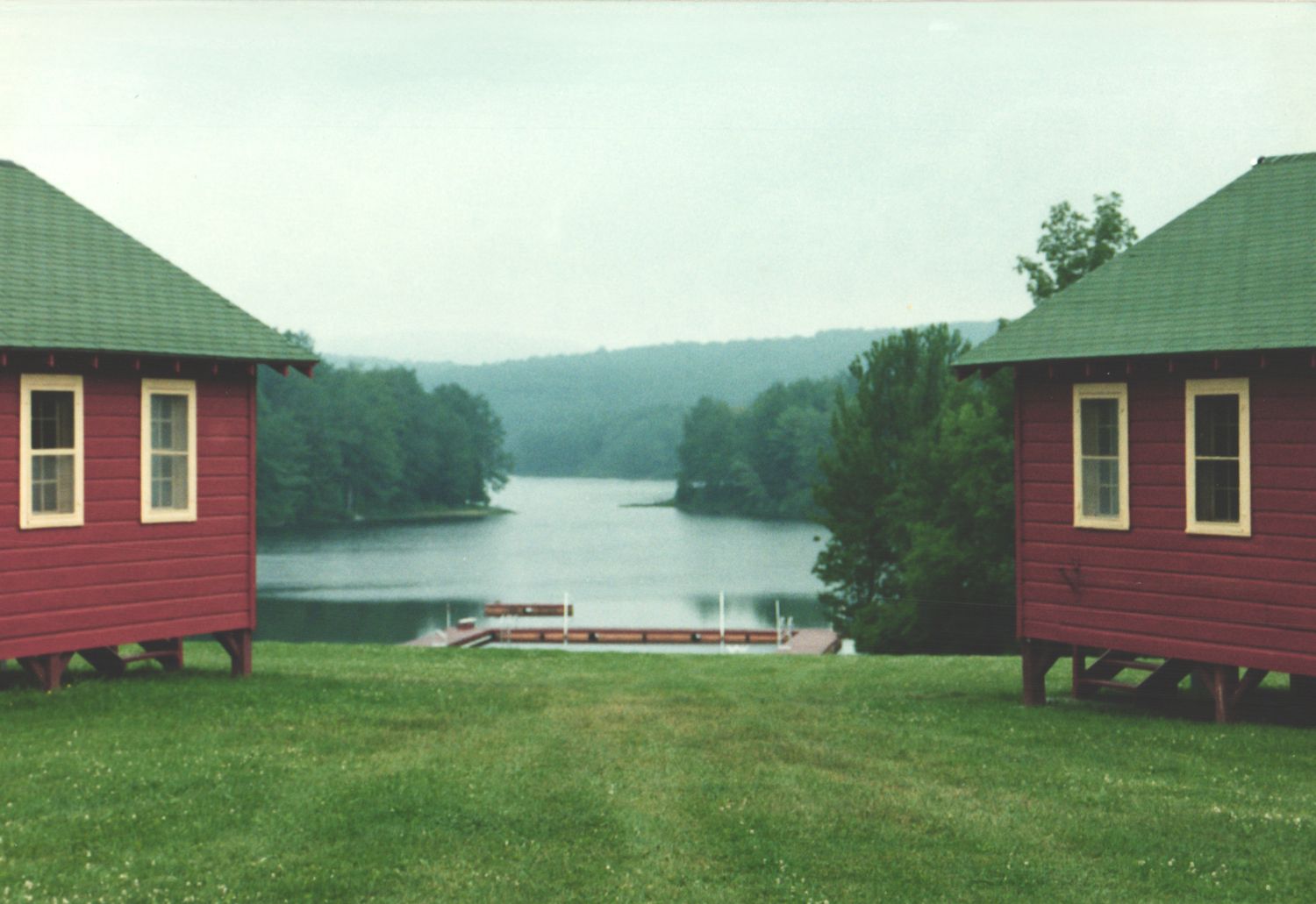 Two red cabins with green roofs framing a calm lake with dock on overcast day.