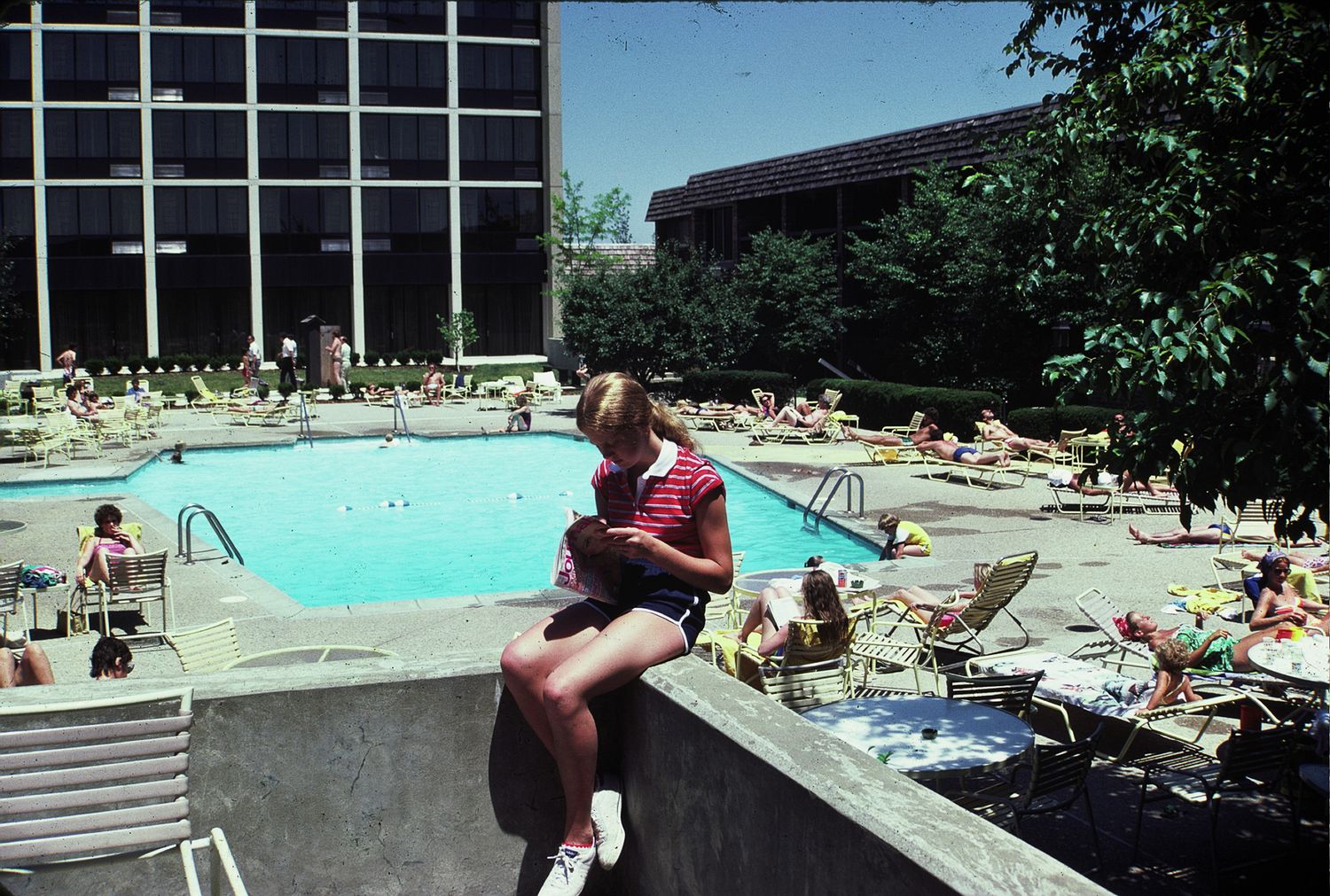 Woman sitting on pool edge reading book at busy outdoor swimming pool surrounded by sunbathers.