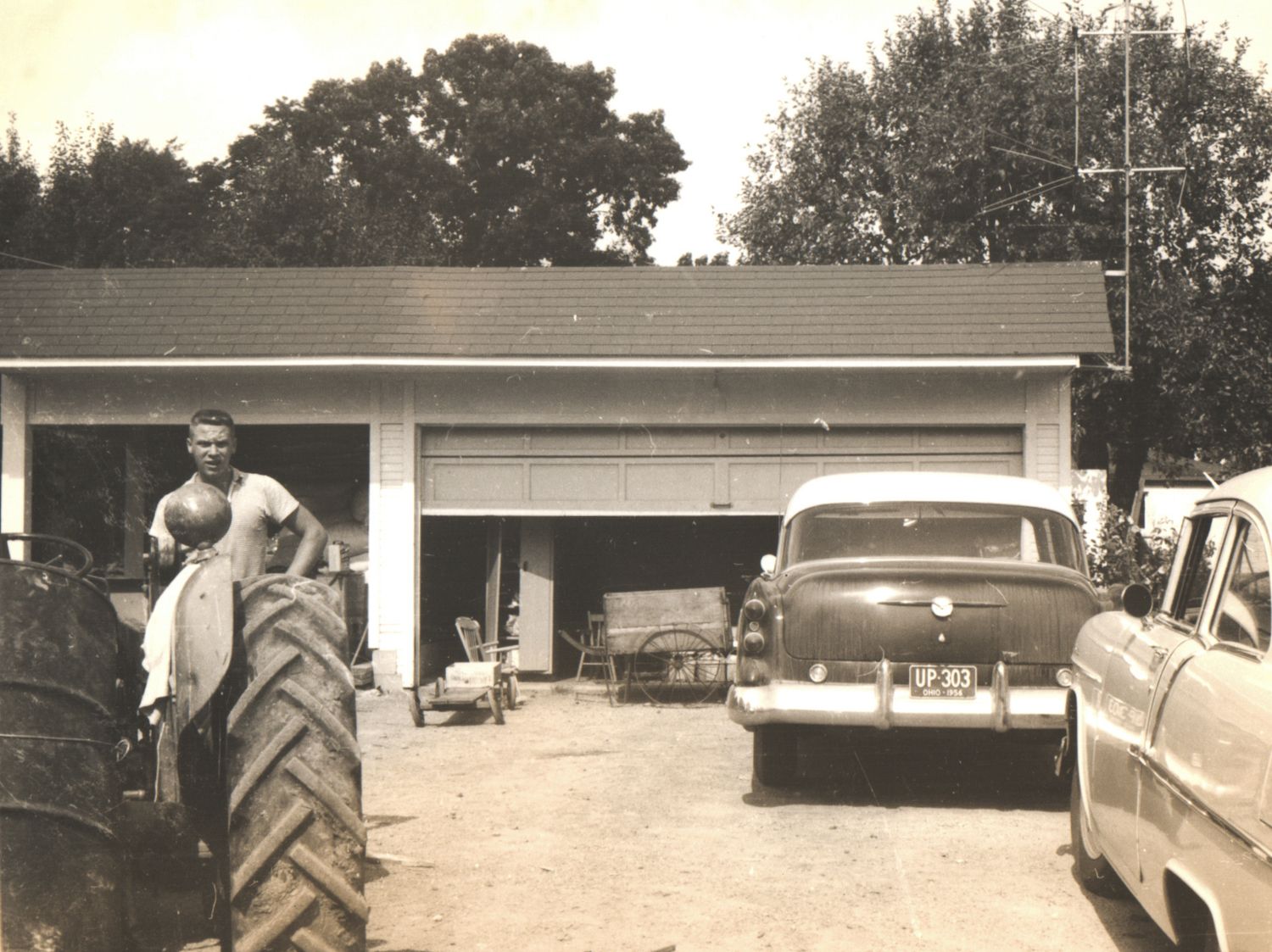 Vintage black and white photo of a man near a large tractor tire outside a rural garage with classic 1950s cars.