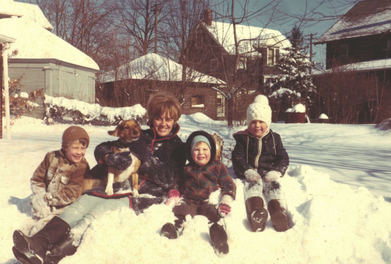 Family with dog posing in snowy backyard during winter, children and woman smiling in deep snow near houses.