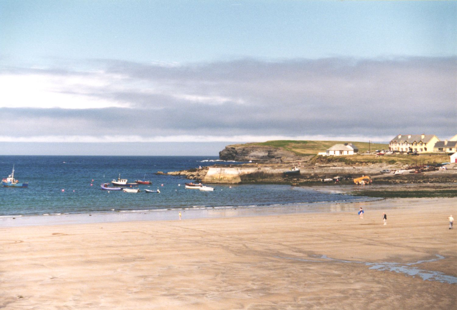 Scenic sandy beach with calm turquoise waters, moored boats, and white coastal houses under a cloudy sky.