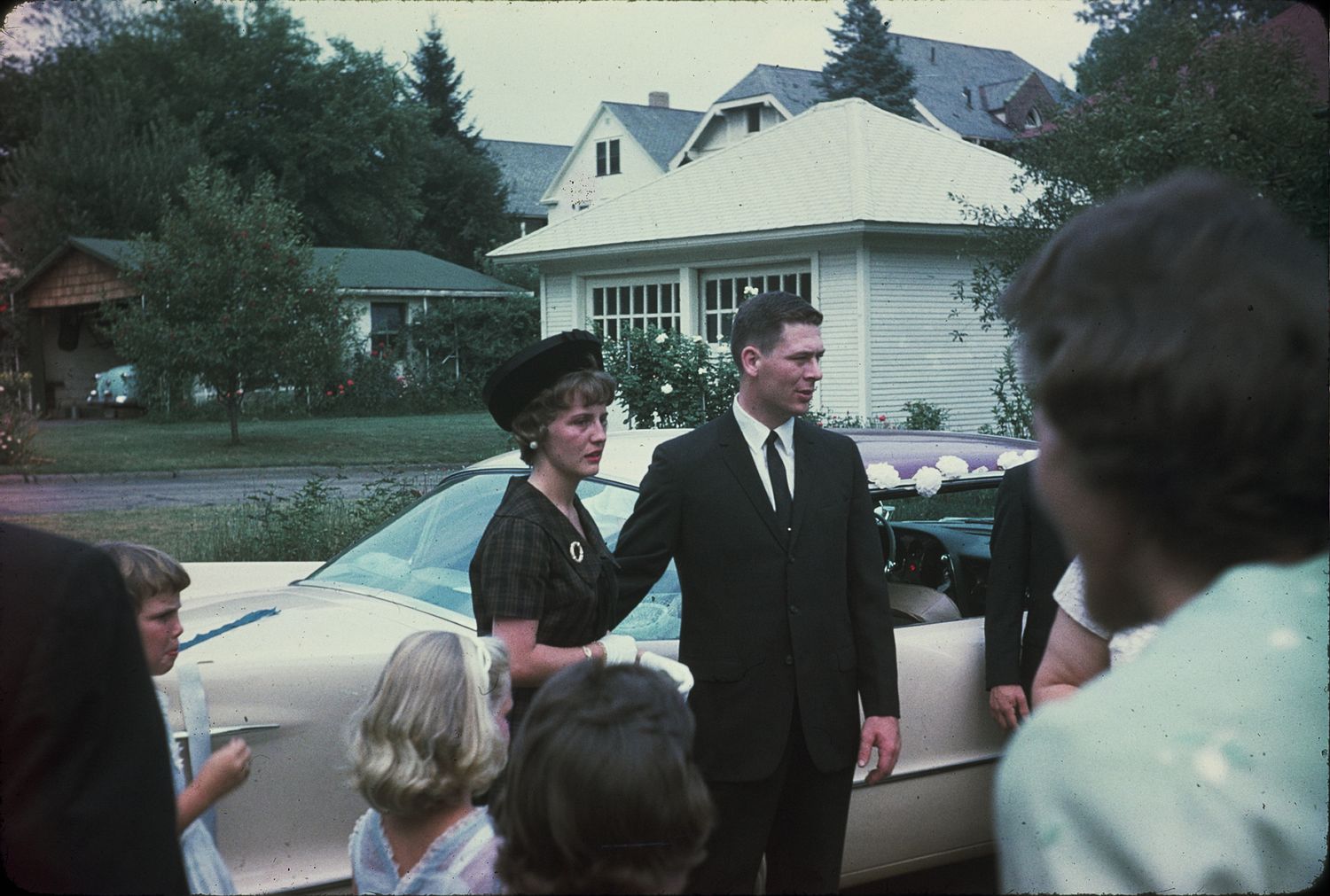 A well-dressed couple shakes hands outdoors near a white car, surrounded by guests at a 1960s suburban gathering.