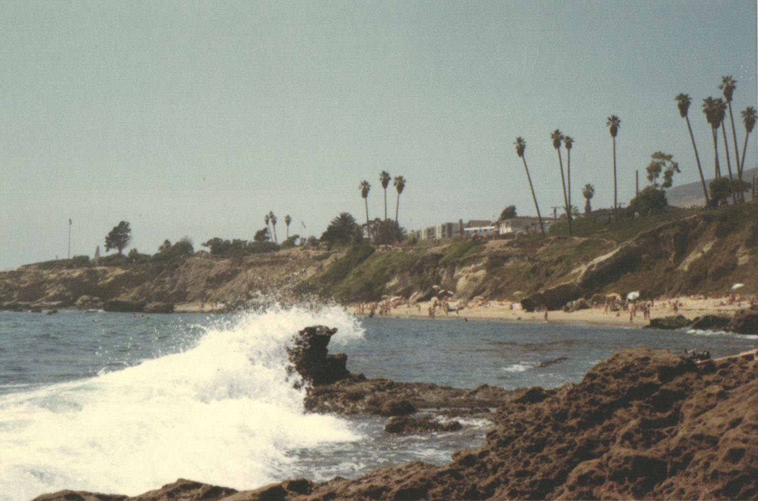 Waves crash against rocky shore with sandy beach, palm trees, and cliffside buildings in the background.
