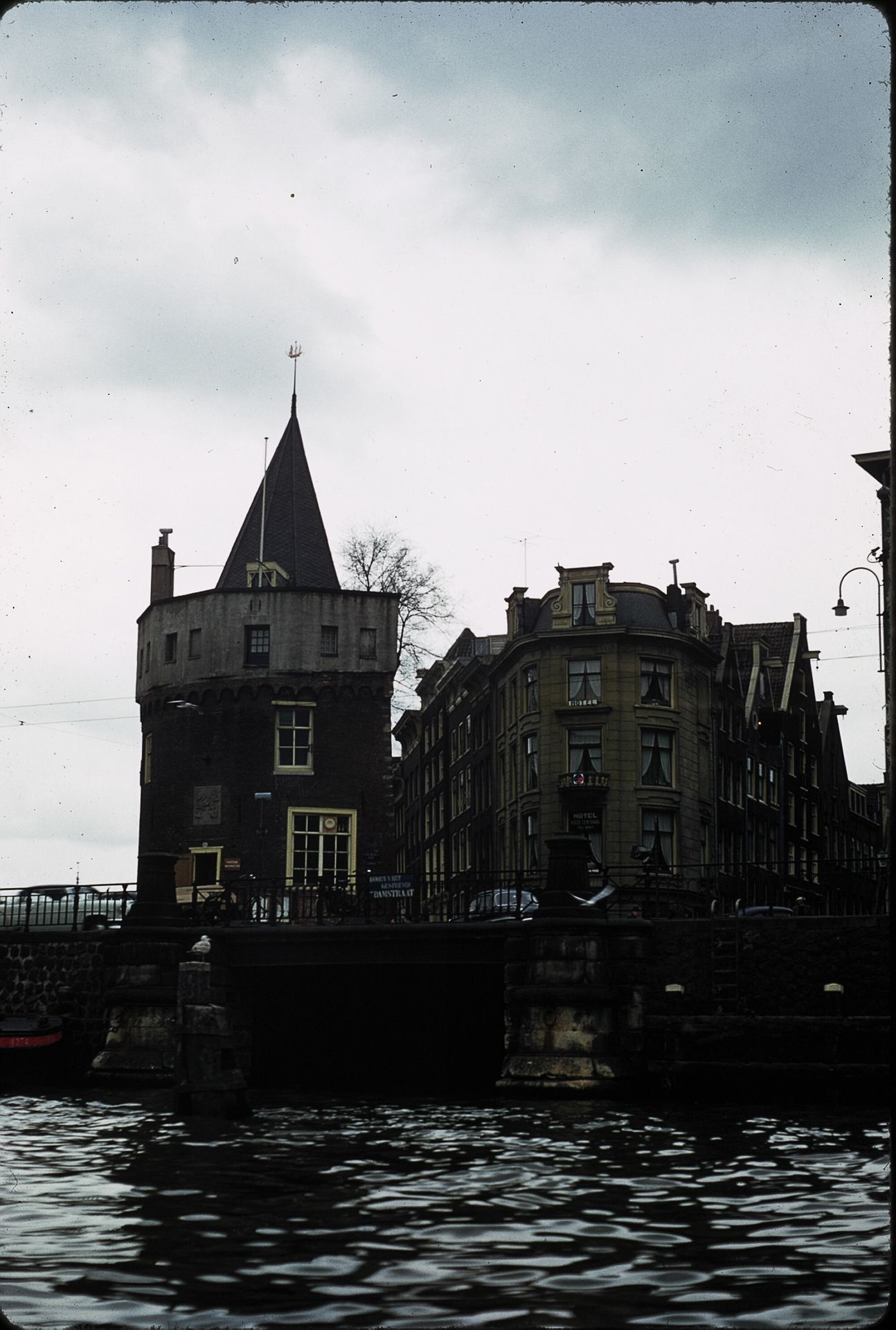 Historic medieval tower and canal buildings reflected in dark water under overcast skies in Amsterdam.