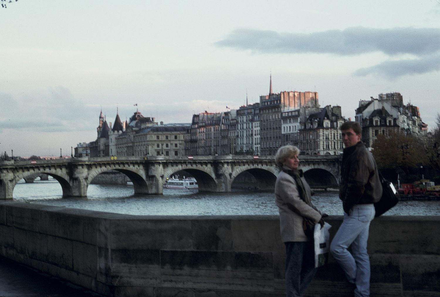 Two people stand along the Seine River in Paris with Pont Neuf bridge and historic cityscape in the background.