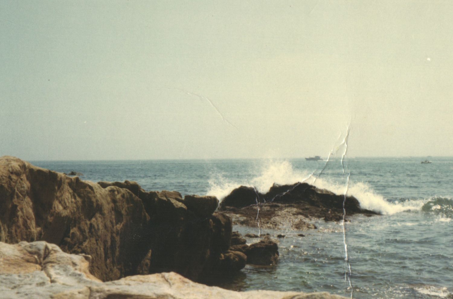 Rocky coastline with ocean waves crashing against dark rocks under a hazy sky.