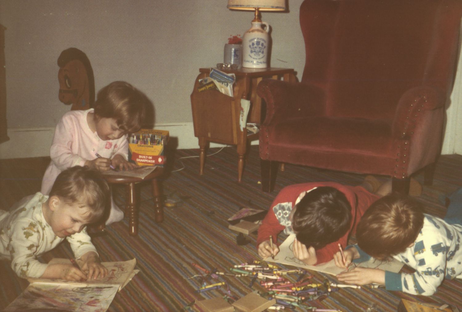 Children lying on a striped rug drawing with crayons in a cozy vintage living room with a red armchair.