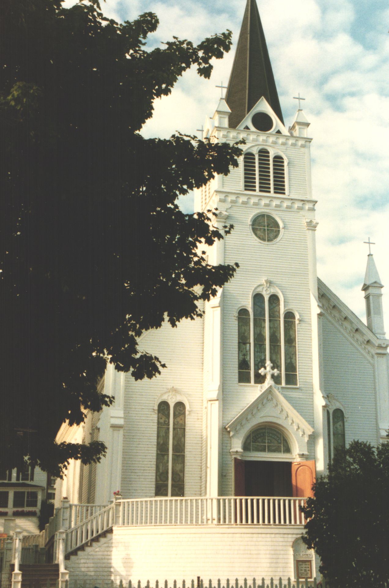 Historic white wooden church with tall steeple, arched windows, and red doors framed by trees against a cloudy sky.