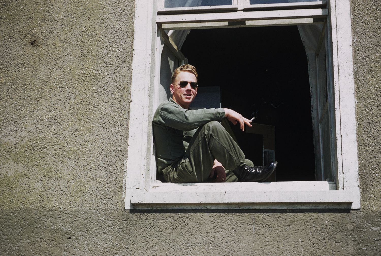 Man in military uniform and sunglasses sitting in open window of gray stone building on sunny day.