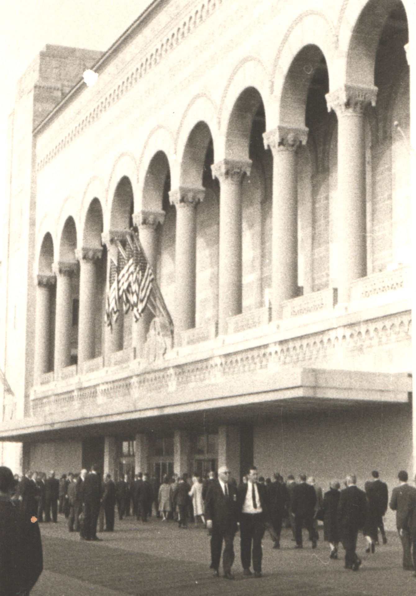 Crowds gather outside a grand arched building facade with American flag in vintage black and white photo.