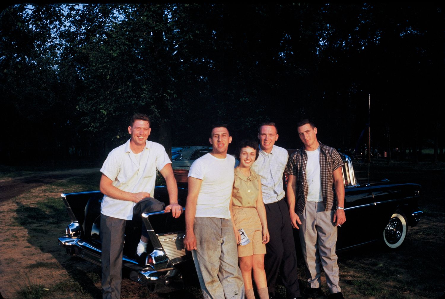 Five young people posing in front of a classic black car outdoors in the 1950s.
