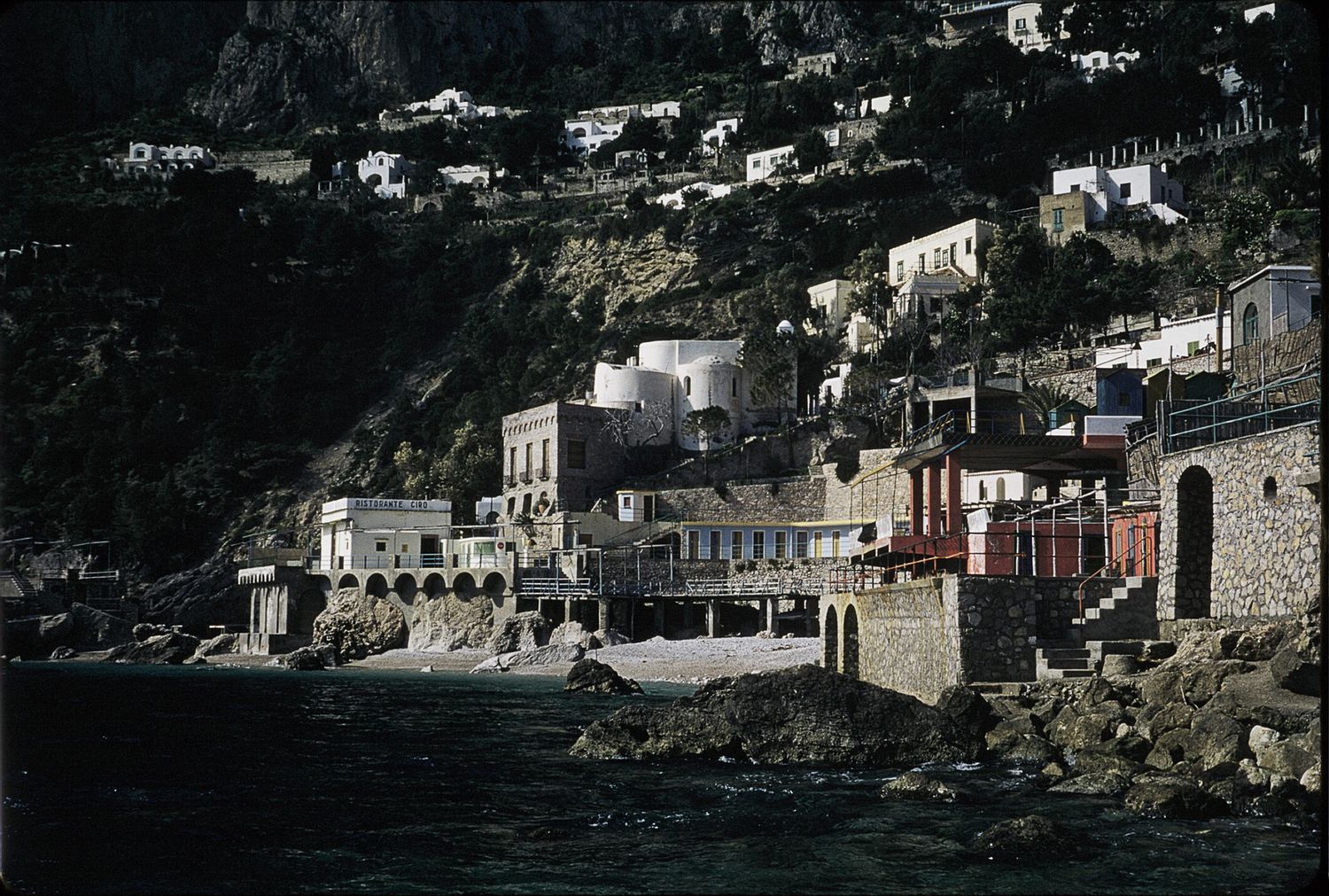 Cliffside Mediterranean coastal village with white buildings, arched facades, and rocky shoreline by the sea.