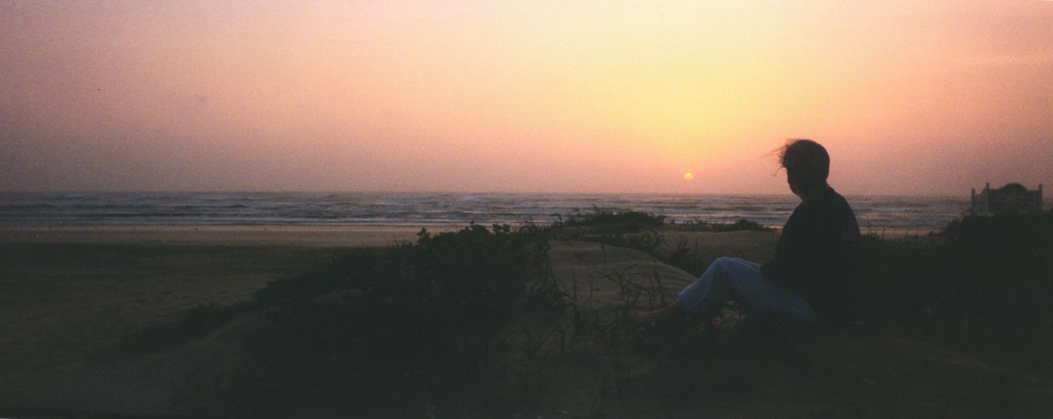 A lone person sits silhouetted on a hilltop watching a breathtaking golden sunset over a vast open landscape.