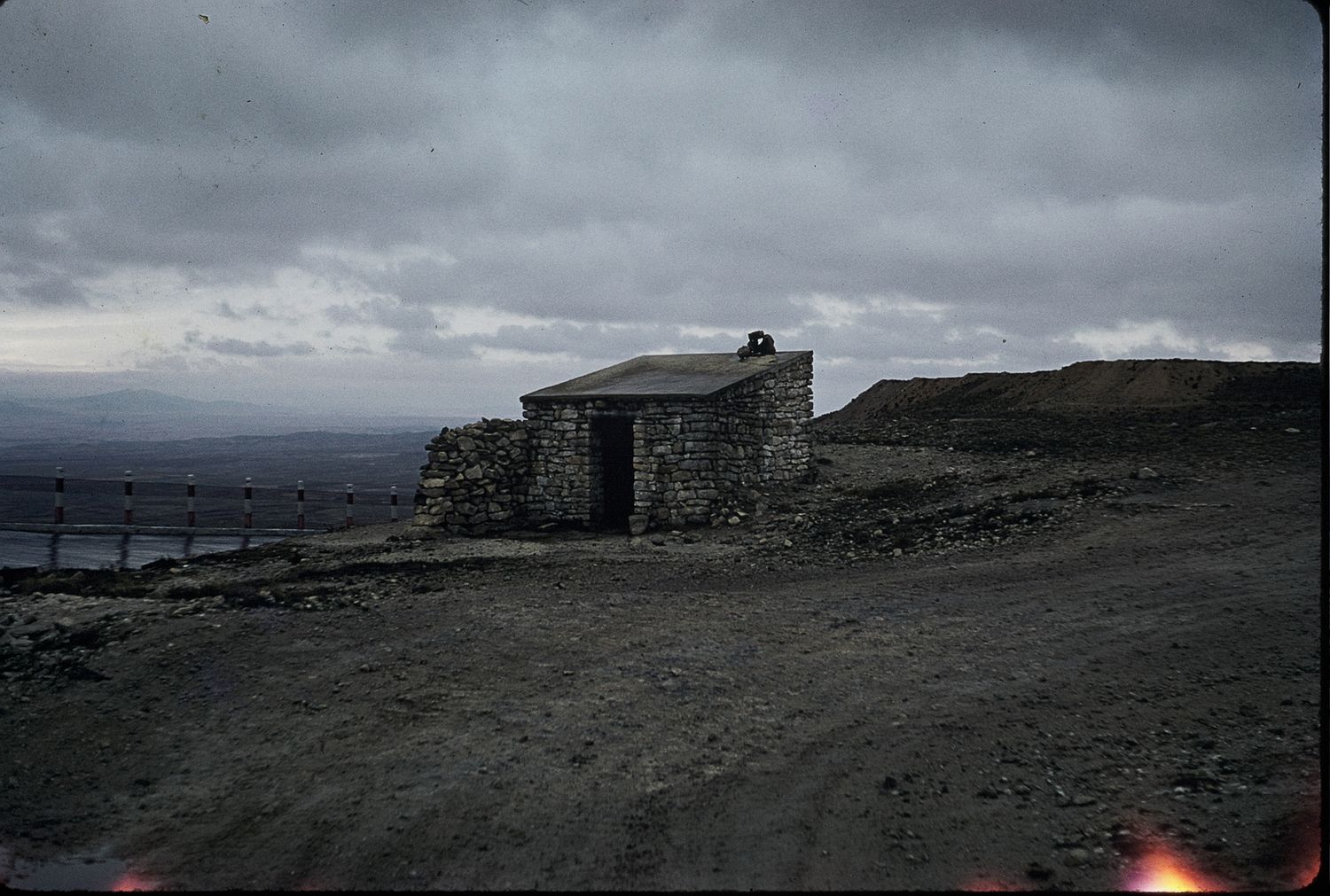 A rustic stone shelter building on a barren highland moorland under dramatic overcast skies with distant hills.