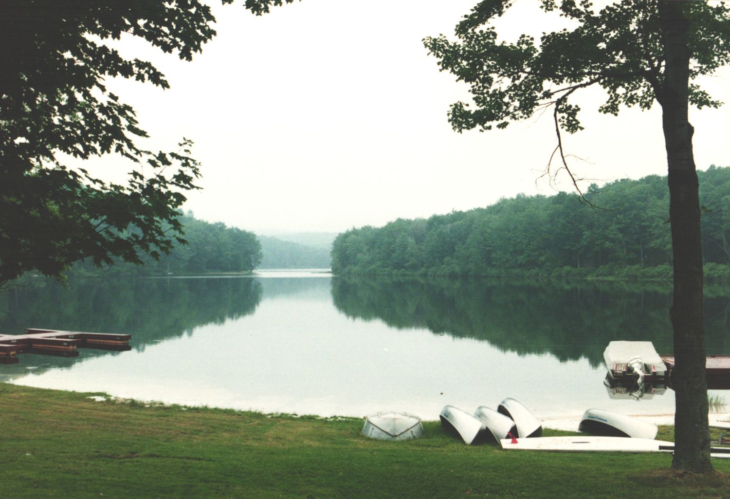Calm lake reflecting trees on a misty morning with canoes resting on the grassy shoreline.