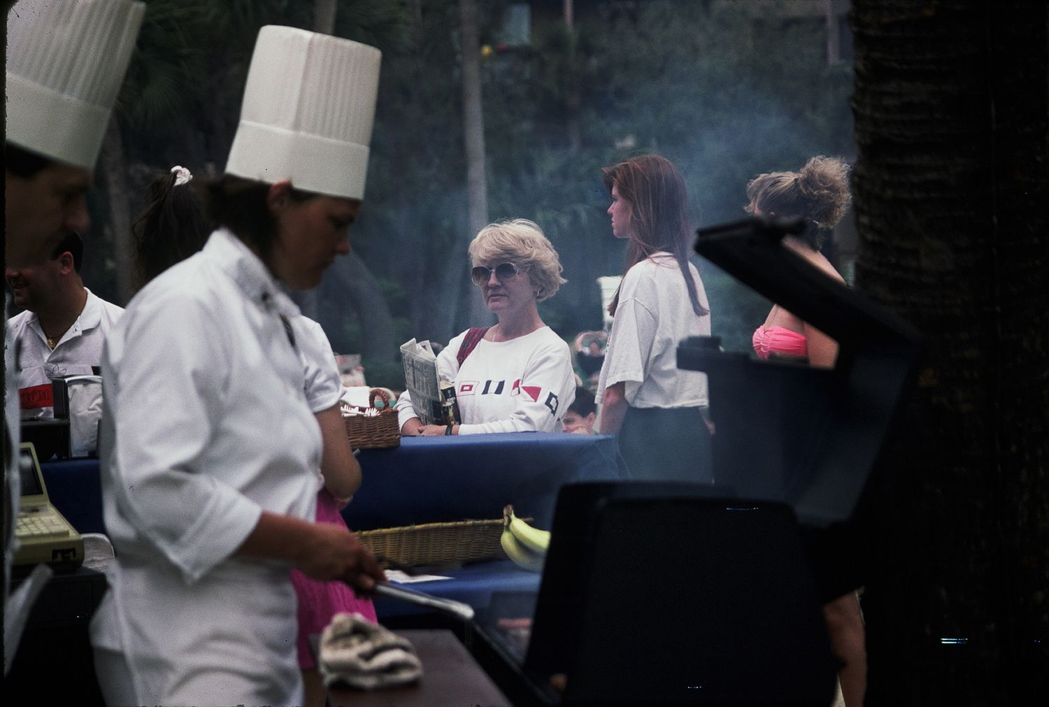 A chef in a white hat grills food outdoors while guests in casual attire gather around a smoky BBQ at an outdoor event.