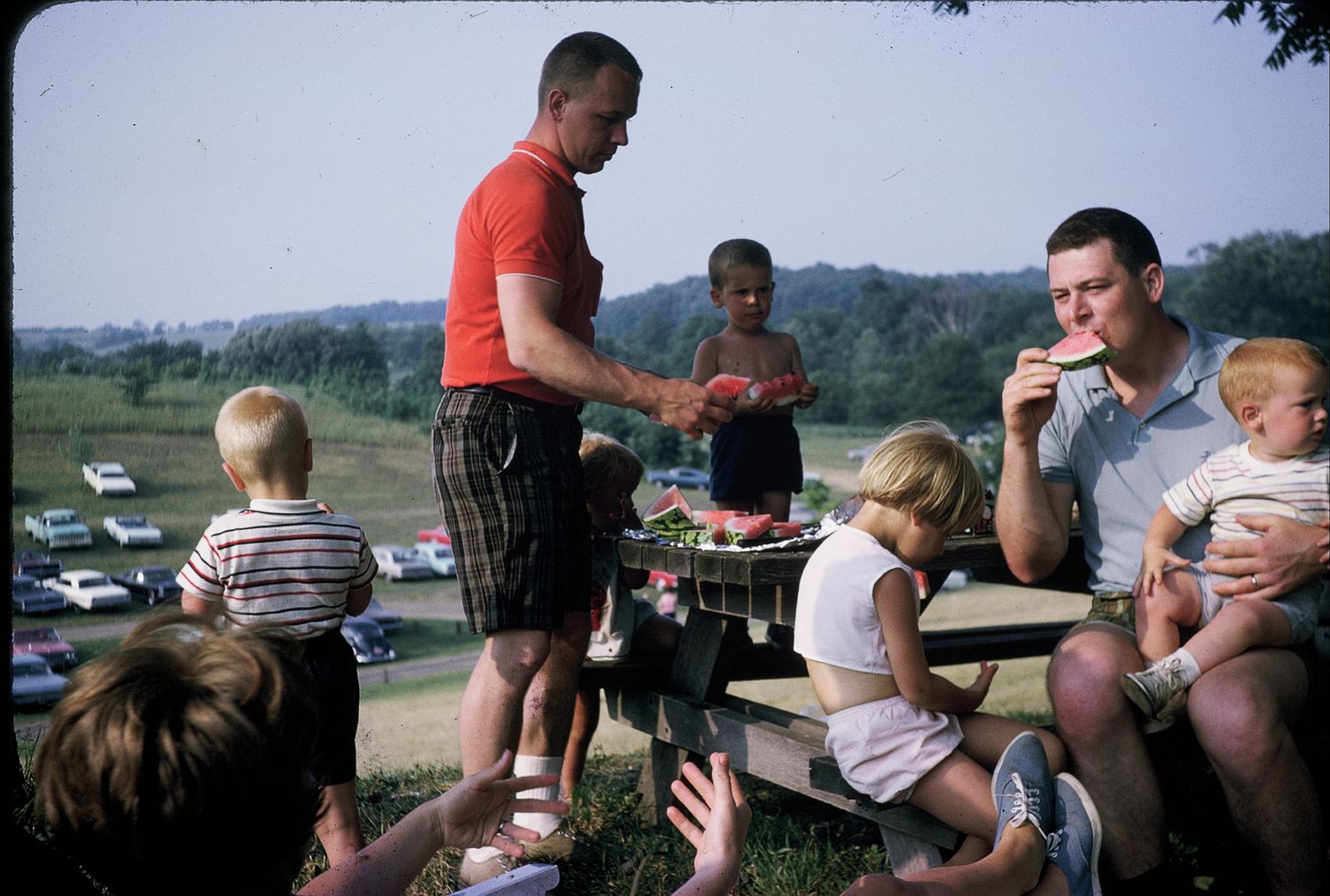 Family outdoor picnic with adults and children sharing watermelon at a wooden table in a green park.