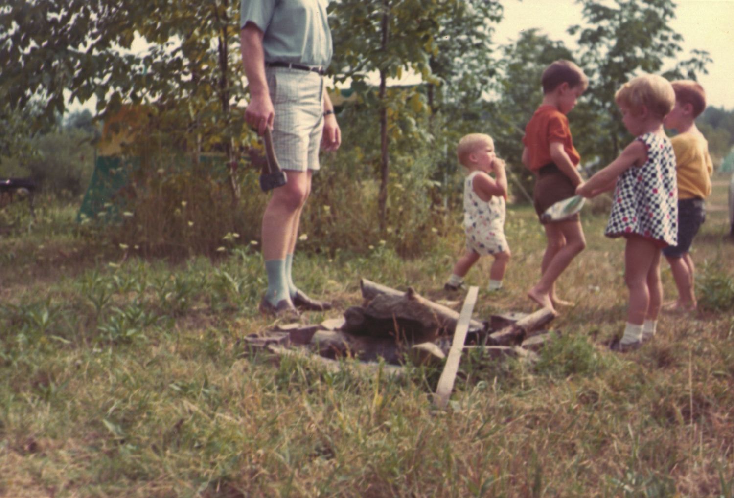 Children playing around a campfire pit in a grassy field supervised by an adult.