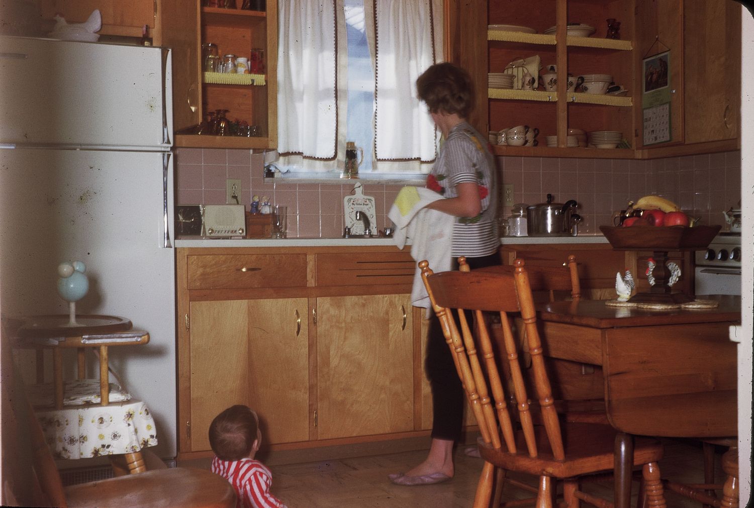 Vintage kitchen scene with woman at sink, wooden cabinets, retro fridge, and toddler playing on floor.