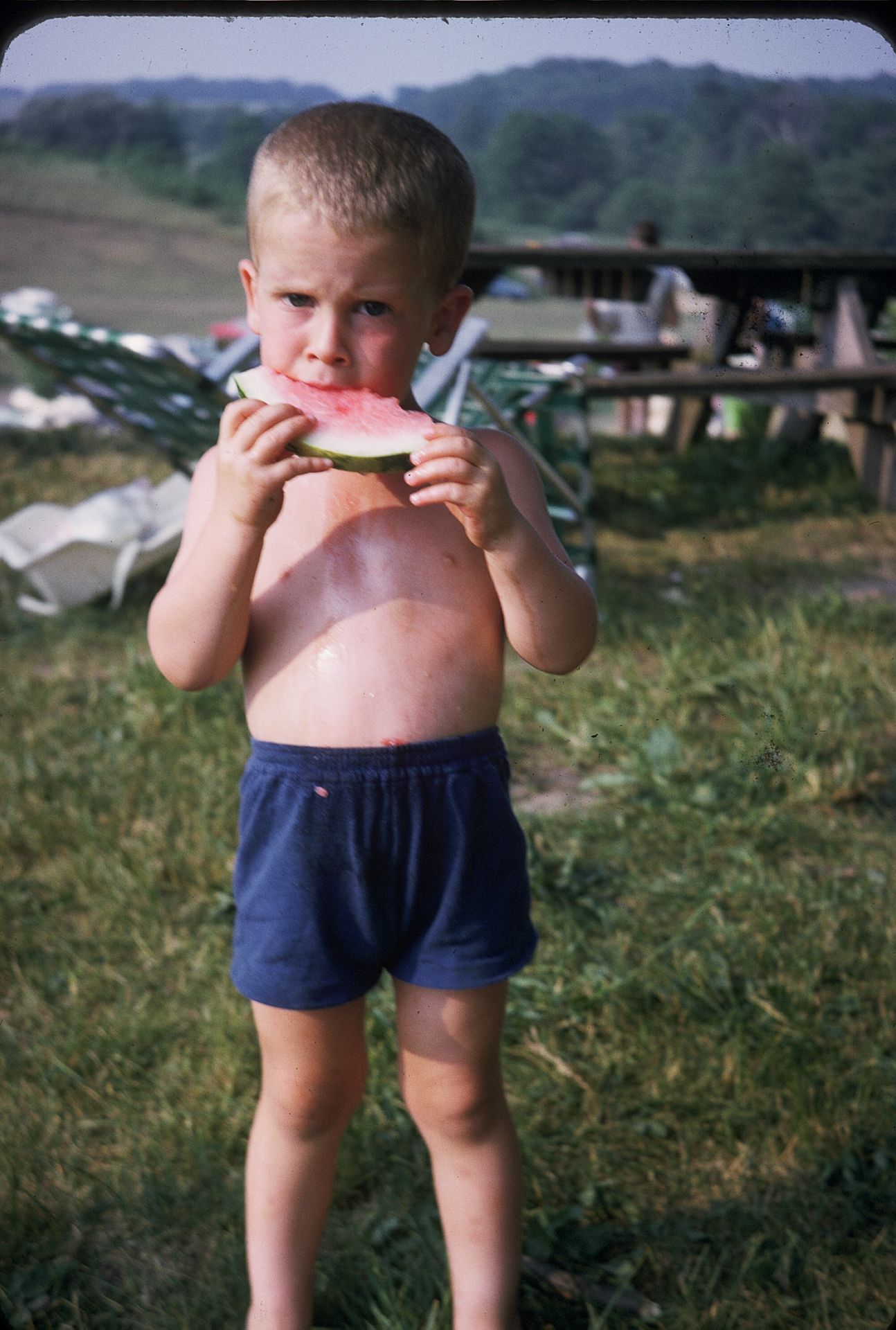 Shirtless young boy eating a fresh slice of watermelon outdoors on a sunny summer day.