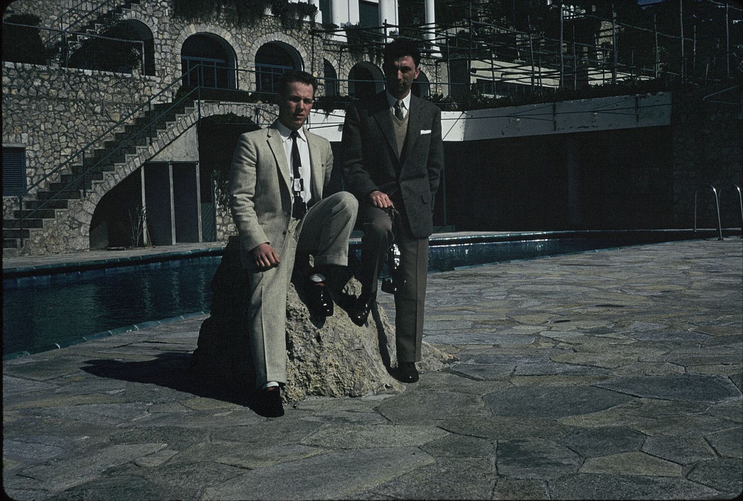 Two men in suits posing by a pool on a stone patio near an ornate building.