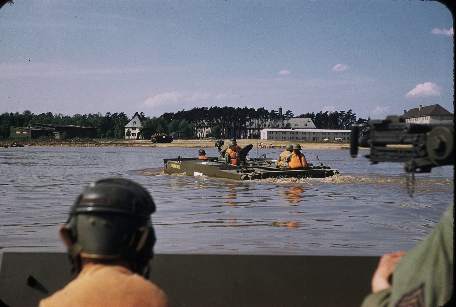 Military personnel navigate a small boat through flooded waters near buildings and trees during a rescue operation.
