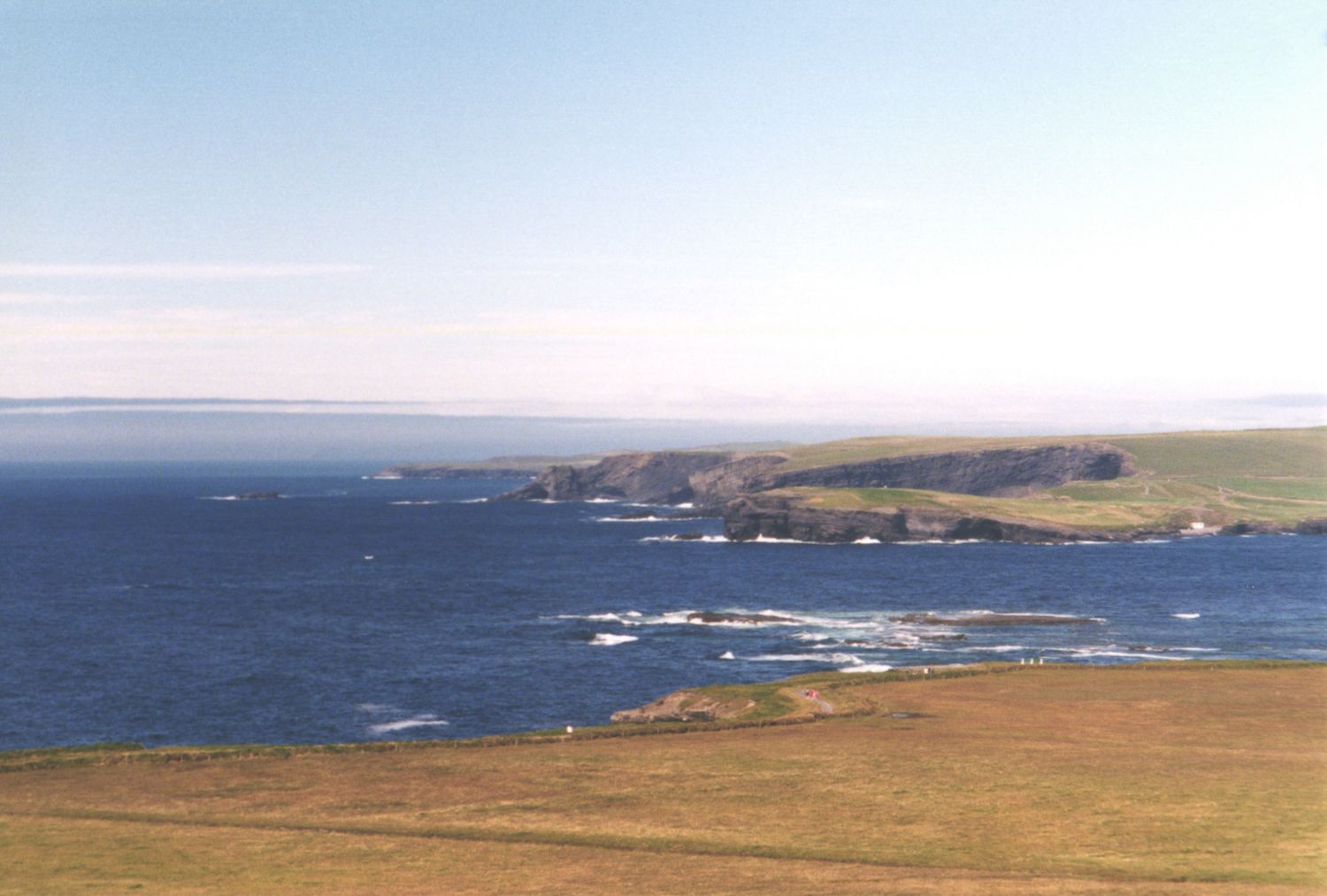 A sweeping aerial view of a rocky coastline with green cliffs meeting the deep blue ocean waters.