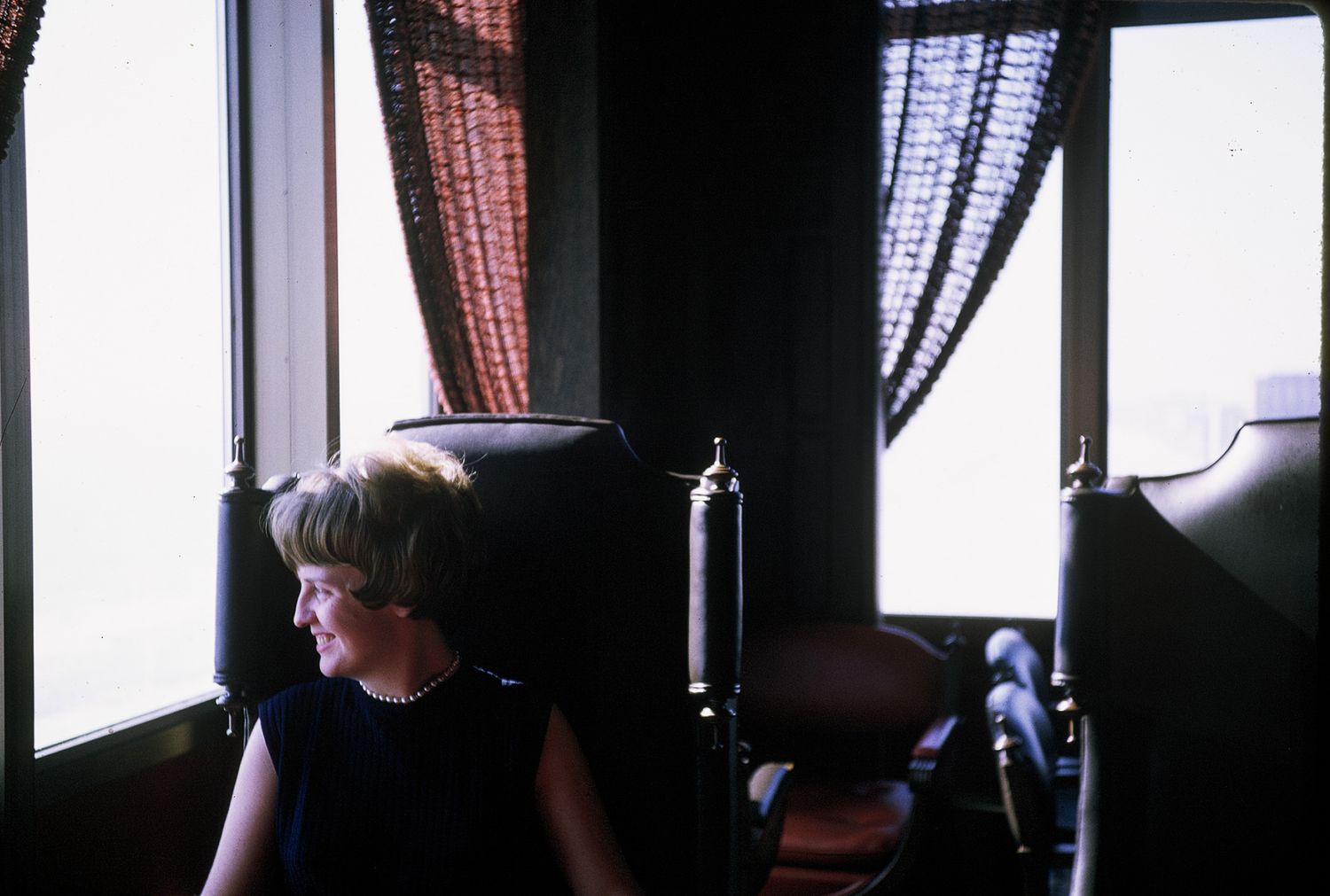 Woman with short blonde hair sits in a chair by a window with dark curtains in a dimly lit room.