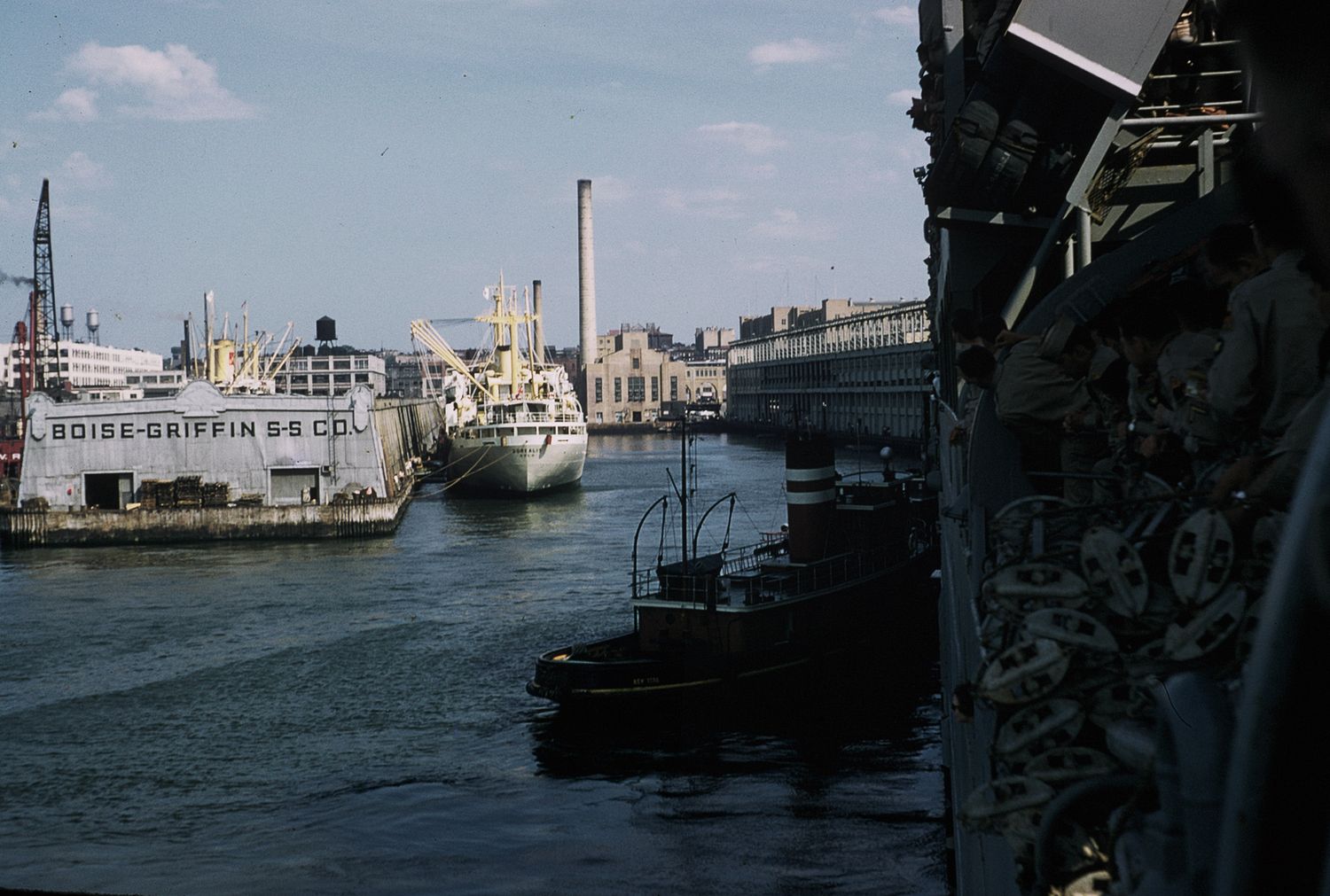 Historic harbor view with docked ships and industrial buildings along a busy waterway on a cloudy day.