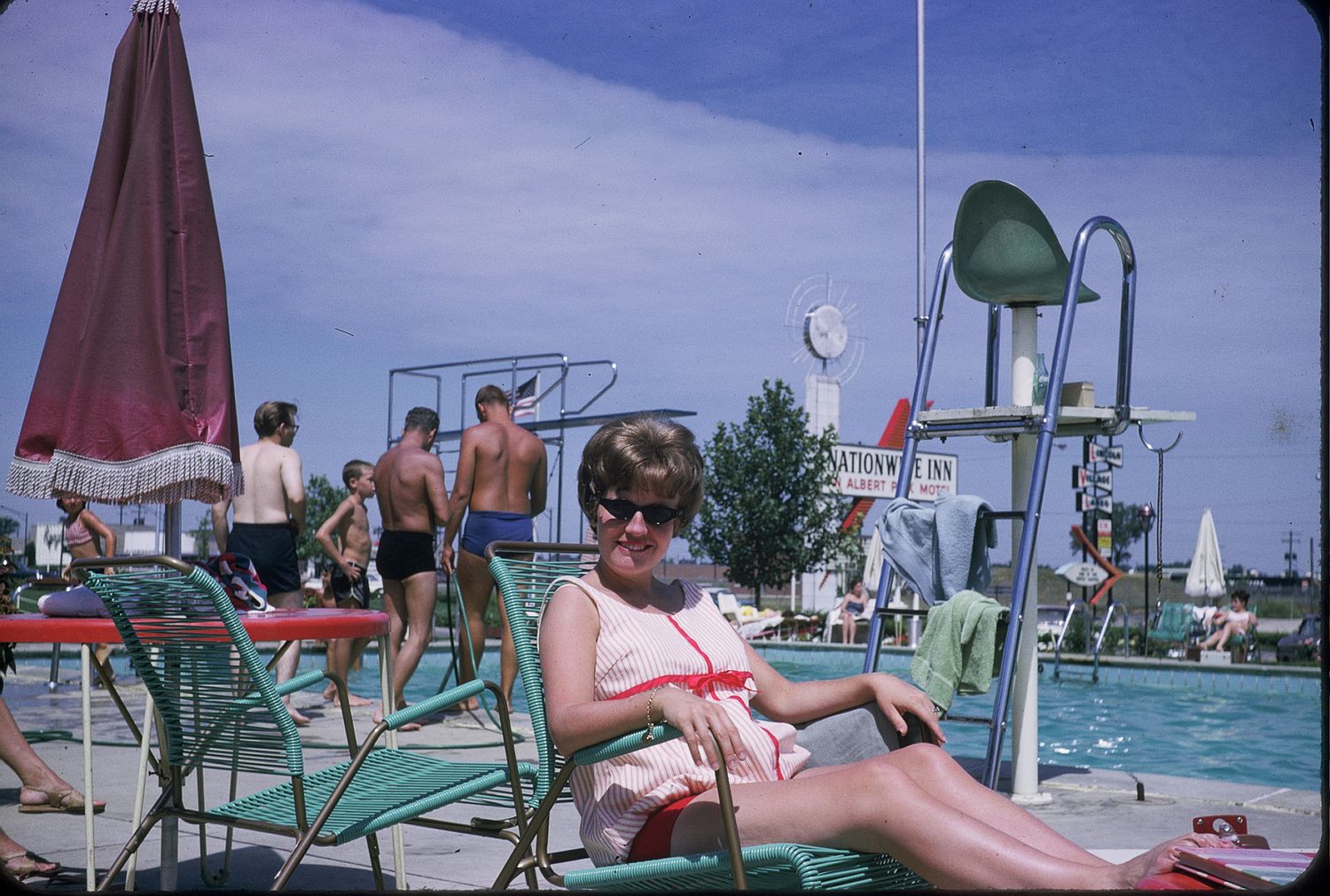 Woman in sunglasses relaxing on lounge chair by outdoor pool with diving board and umbrella on sunny day.