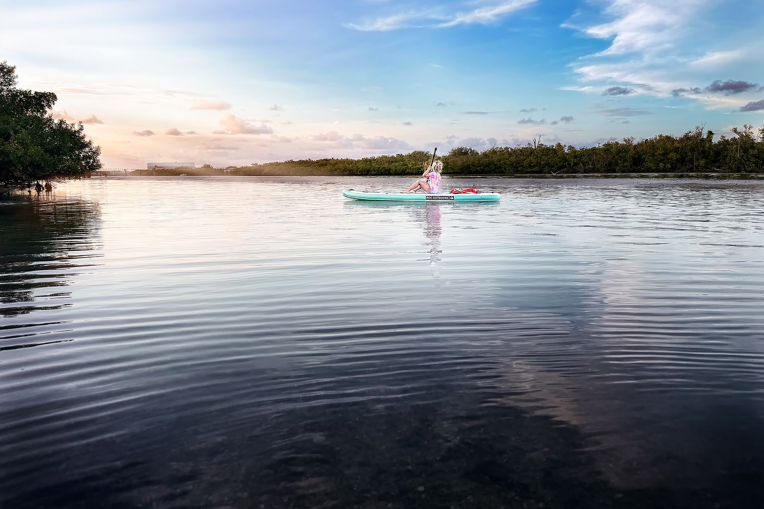 A person kayaks peacefully on calm reflective waters surrounded by mangroves at a serene sunset.