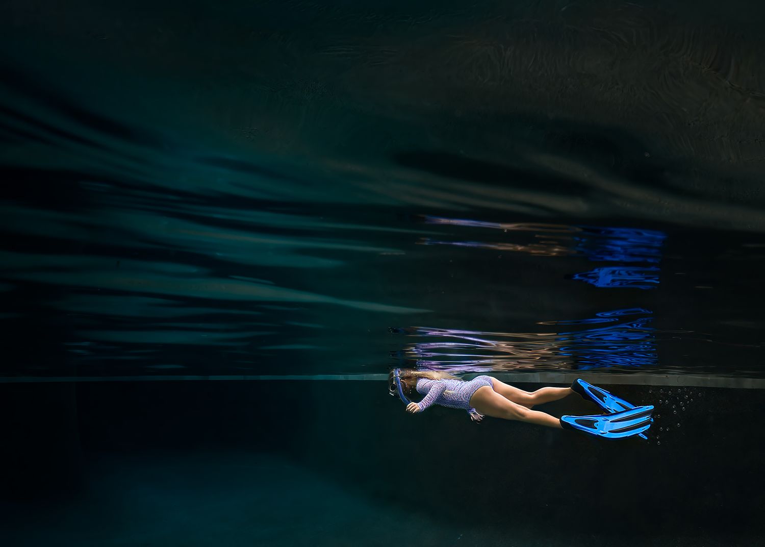 Underwater photo of a child snorkeling with blue fins in dark, serene turquoise pool water.