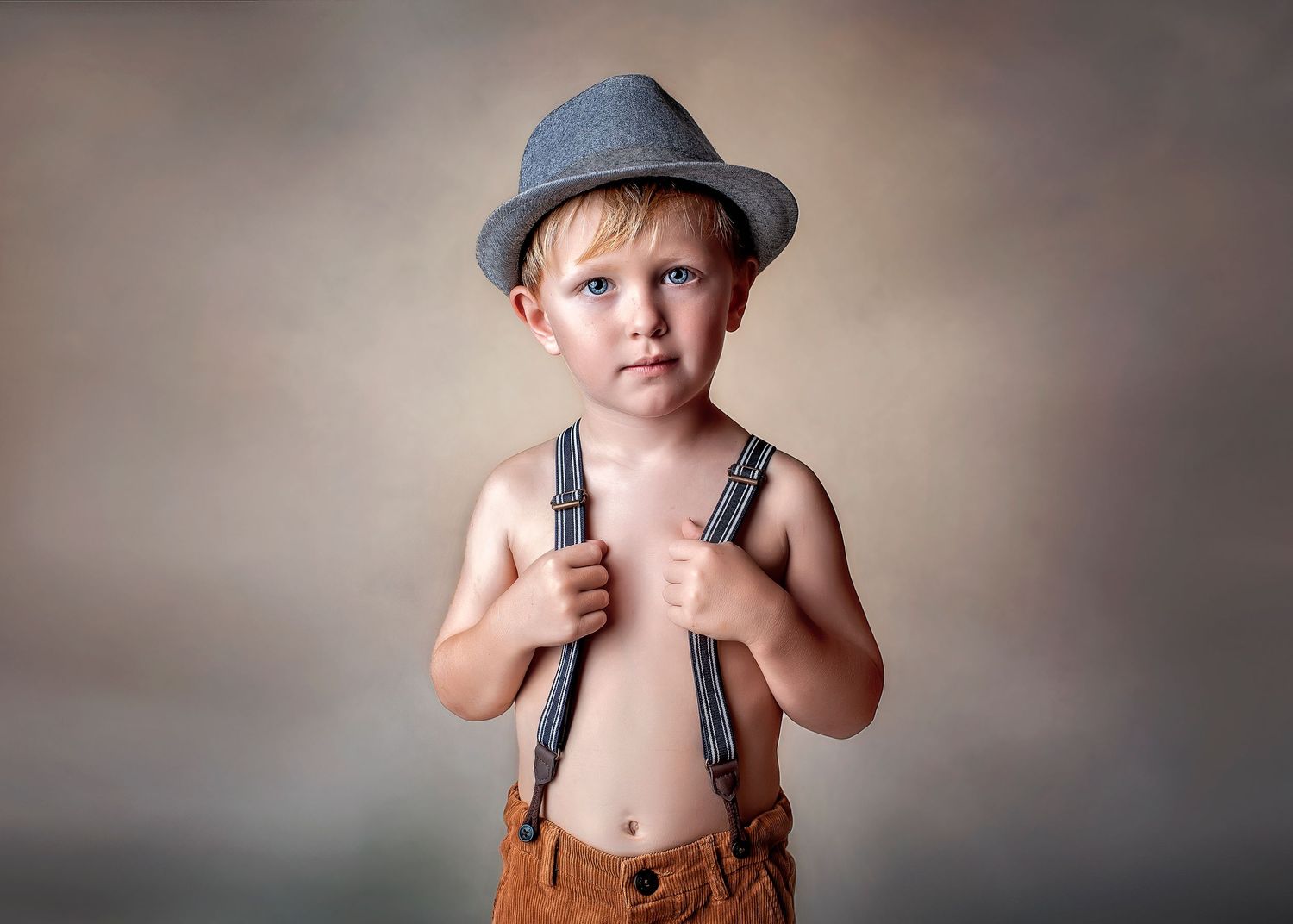 Cute toddler boy wearing hat and suspenders posing for a studio portrait.