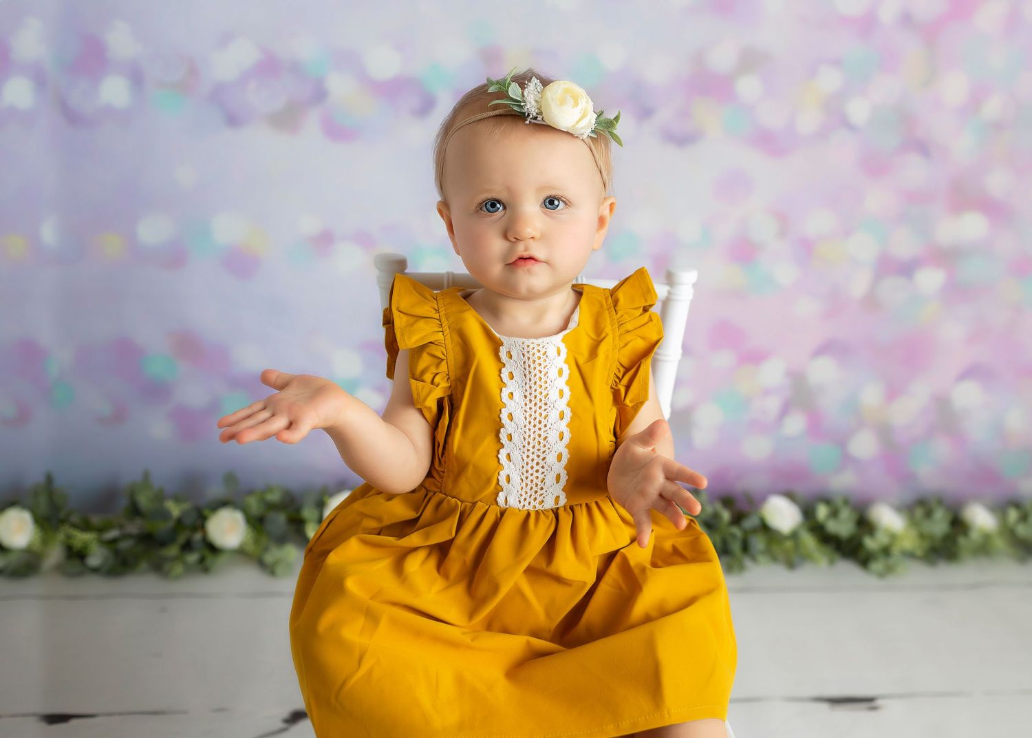 Baby girl in yellow dress with flower crown sitting on white chair against bokeh backdrop.